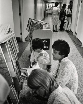 Picture of a librarian crouched on a hospital hallway floor showing a book to a small girl with…