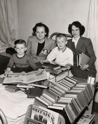 Picture of a two librarians sitting with two boys on a hospital bed looking at a pile of books …