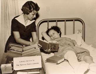 Picture of a librarian showing a book to a child in a cast lying in a hospital bed. 