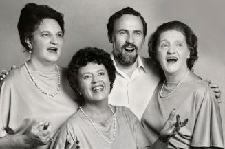 Three women in cape silky sleeved tops and pearls sing with a bearded man in between in a white…