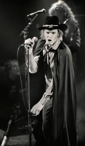 On an darkened stage, a man in confederate hat and black cape and patterned shirt twists his mo…