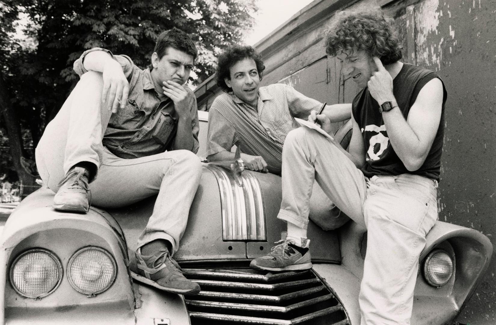 three young men sit and lean on the hood of an old 1950s car, one is stroking his chin and anot…