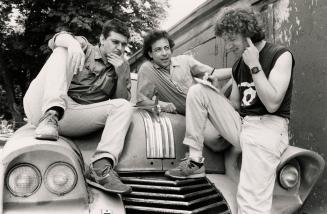 three young men sit and lean on the hood of an old 1950s car, one is stroking his chin and anot…