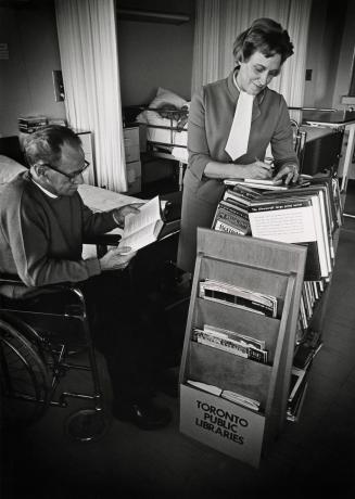 Picture of a librarian with library truck of books showing books to a man in a wheelchair in a …