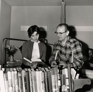 Picture of a young woman showing a book from a book truck to a man in a hospital room. 