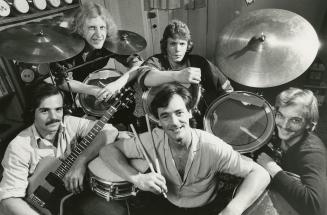 5 young men surround a large drum kit. They wearing shirts and have shaggy hair. One on the low…
