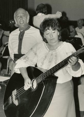 A woman is playing a very large mandolin and over her right shoulder is a man who is singing. T…