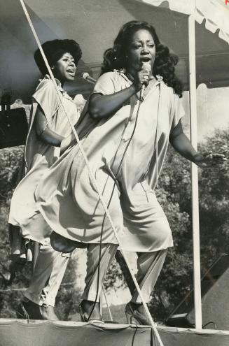 Two ladies in heels and flowy pajama dresses sing on stage in the sunlight