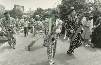 On the street daytime, 3 men in loud, patterned suits parade with their saxophones, followed by…