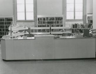 Picture of a 1960 library desk with shelves behind and two large windows.
