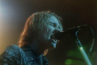 Young man with light coloured shaggy hair singing into microphone on stand and playing guitar