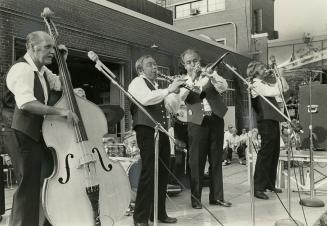 4 middle aged men in matching trousers and vests behind microphones on stands play front left t…