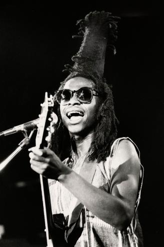 Performing live with guitar and microphone, man in sunglasses with a tall black braided cone on…
