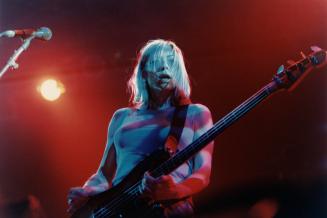 Young woman with shaggy blond hair in t-shirt in playing bass guitar with microphone on the lef…