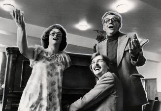 Around an upright piano, a woman singing, a man sitting and playing piano and another man singi…