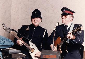 Two policemen in uniform play guitars and sing into a shared microphone. White wallpaper backgr…