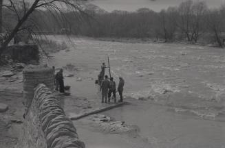 Photograph of a several people fishing in a river with stone wall in foreground. 
