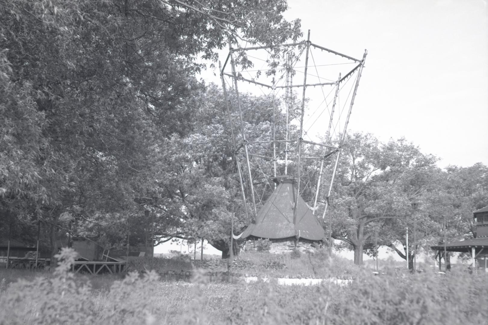 Photograph of an abandoned wooden amusement park ride with overgrown trees on right and grasses…