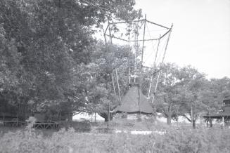 Photograph of an abandoned wooden amusement park ride with overgrown trees on right and grasses…