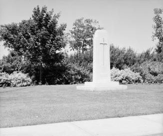 Photograph of a stone memorial with a cross surrounded by lawn and bushes and trees behind. 