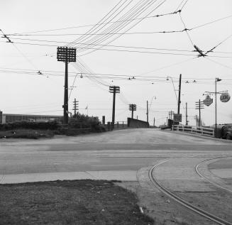 Photograph of intersection showing a bridge, streetcar lines and several hydro poles and wires.…