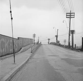 Photograph of a road heading over a bridge showing a wall with large writing on the left and se…