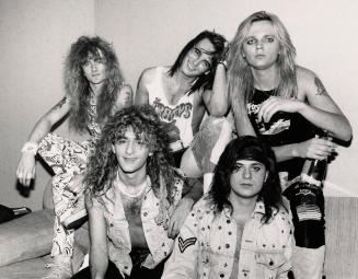 5 young men clumped tonight on a sofa against a white wall. They have long wavy hair and wear t…