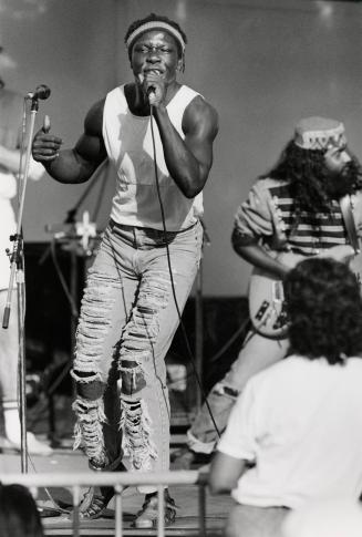 On stage, man in tank top and ripped jeans sings into hand held microphone. Guitar player to hi…