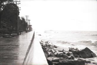 Photograph of a wooden boardwalk with rocks and waves from lake on right and trees and glimpse …