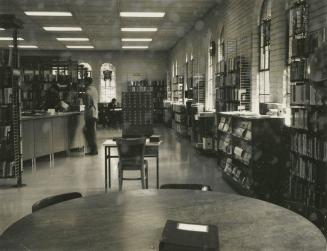Photograph of interior of a library with stained glass windows, table in foreground, shelves ro…