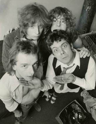 Four young men in a tight cluster look up into the camera, with shaggy hair and shirts on. 