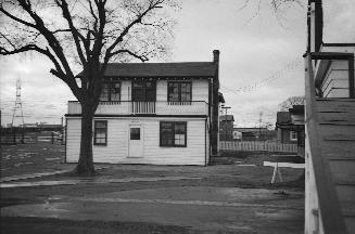 Photograph of a small two storey house with tree in front and some wooden building partially se…