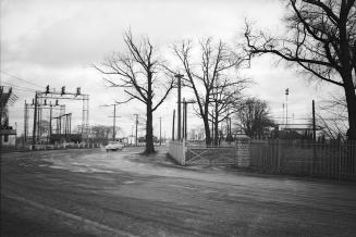 Photograph of a road with gate on right side and bare trees and on left some hydro poles and wi…