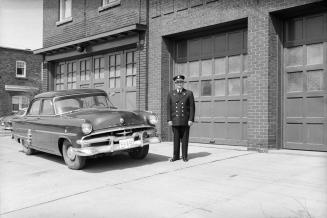 Photograph of man in fire department uniform standing beside a car in front of fire station dou…