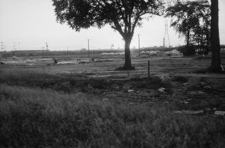 Photograph of a field with a few trees in foreground and some building materials and cars and t…