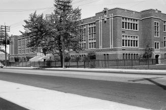 Photograph of corner view of three storey brick school with tree in front. 