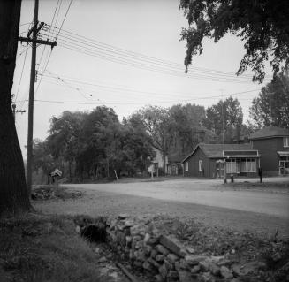 Photograph of dirt road intersection with gully and drain in foreground and houses in backgroun…