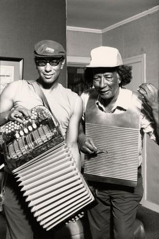 Two men, on left with accordion, on the right with a washboard. Casually dressed with caps. 
