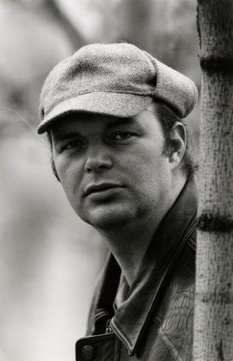 Man in cap beside tree wearing leather jacket. short hair, clean shaven