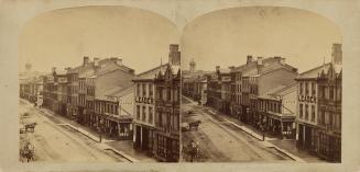 Picture looking down a dirt street with buildings on one side, wooden pavements and two horses.…