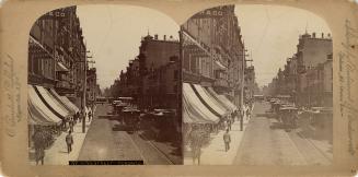 Picture of a street with buildings on both sides and shops with large awnings and streetcars go…