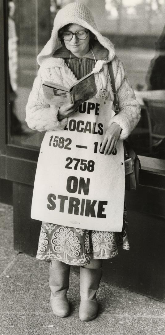 Middle-aged woman with glasses in parka reads a book wearing banner saying, 'CUPE locals...on s…
