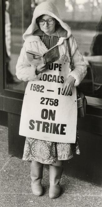 Middle-aged woman with glasses in parka reads a book wearing banner saying, 'CUPE locals...on s…