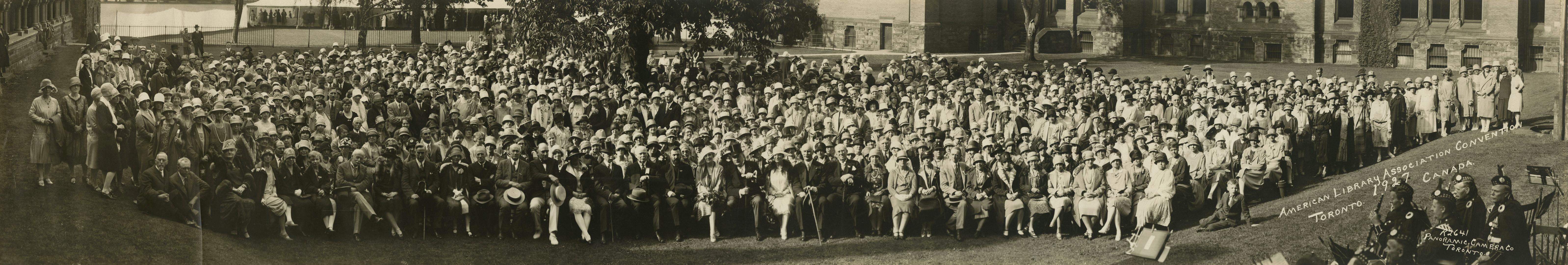 Black-and-white panoramic photograph showing a very large group of people arranged in dense row…