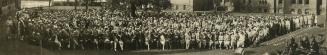 Black-and-white panoramic photograph showing a very large group of people arranged in dense row…
