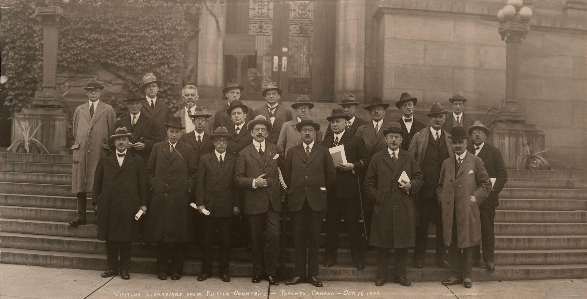 Black-and-white group photograph of about sixteen adults standing on stone steps outside a larg…