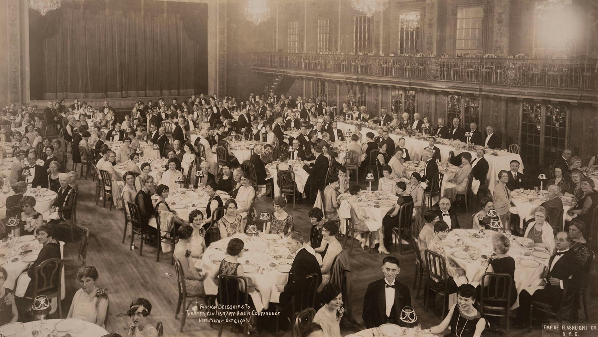 Black‑and‑white photograph of a large formal banquet in a grand hall. Long rows of round tables…