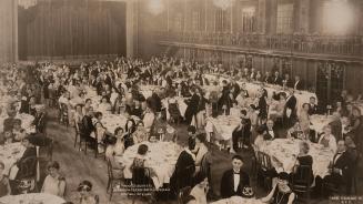 Black‑and‑white photograph of a large formal banquet in a grand hall. Long rows of round tables…