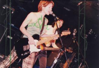 2 young women on dark stage, playing guitars. The first one is singing. . 