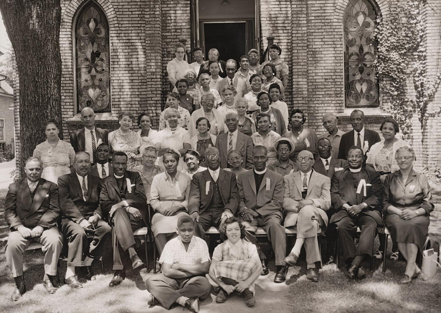Black-and-white photograph of a large group of adults and children gathered in two rows outside…